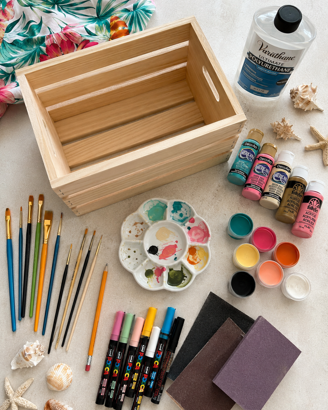 Supplies for painting DIY bikini boxes including a wooden crate, acrylic paint, brushes, sandpaper, and paint markers.